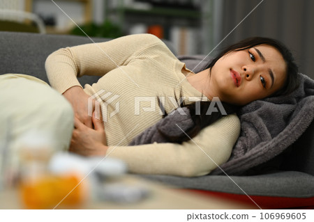 Unhappy woman in painful expression holding her belly suffering from stomachache or menstrual period pain lying on couch Unhappy woman in painful expression holding her belly suffering from stomachache or menstrual period pain lying on couch 106969605
