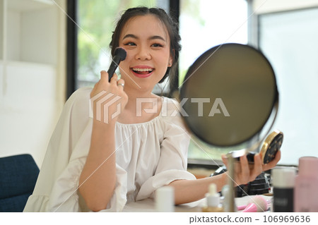Cheerful young woman looking at mirror and applying makeup with brush on cheeks. Beauty, cosmetic and lifestyle concept 106969636