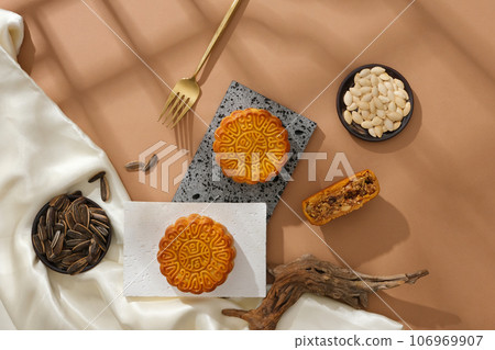 Mid-Autumn Festival traditional food concept - mooncakes and types of seeds decorated with white silk cloth on brown background. Top view, scene for advertising Mid-Autumn Festival traditional food concept - mooncakes and types of seeds decorated with white silk cloth on brown background. Top view, scene for advertising 106969907