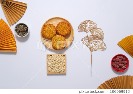 Above view of wooden trays filled with appetizing mooncakes and different types of seeds decorated with yellow paper fan on white background. Background of Chinese Traditional Mid-Autumn Festival Above view of wooden trays filled with appetizing mooncakes and different types of seeds decorated with yellow paper fan on white background. Background of Chinese Traditional Mid-Autumn Festival 106969913