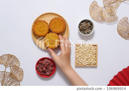 Mooncake advertising scene with modern concept - on white background with some golden decorations, wooden dish of mooncakes displayed with trays of jam and seeds. Hand is holding a mooncake 106969914