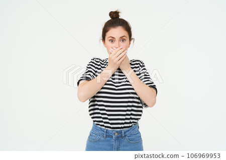 Portrait of woman looking shocked, covers her mouth, looks speechless and amazed, stares with disbalief, stands over white background 106969953