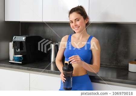 Portrait of fit and healthy smiling woman, wearing fitness sportswear, standing in kitchen with water bottle and looking happy Portrait of fit and healthy smiling woman, wearing fitness sportswear, standing in kitchen with water bottle and looking happy 106970018