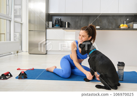 Portrait of beautiful brunette fitness girl, plays with her dog during workout training session, sits at home on rubber yoga mat, does sport exercises in living room Portrait of beautiful brunette fitness girl, plays with her dog during workout training session, sits at home on rubber yoga mat, does sport exercises in living room 106970025