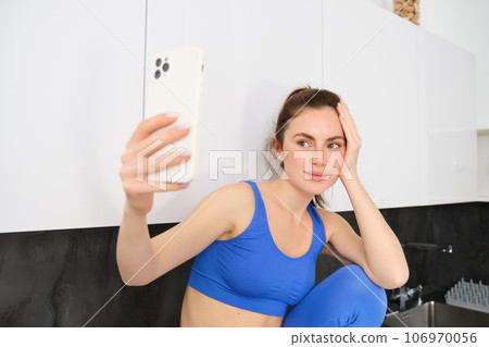 Portrait of beautiful sportswoman, wearing sportsbra and leggings, sitting in kitchen and taking selfie, posing for photo on smartphone app 106970056