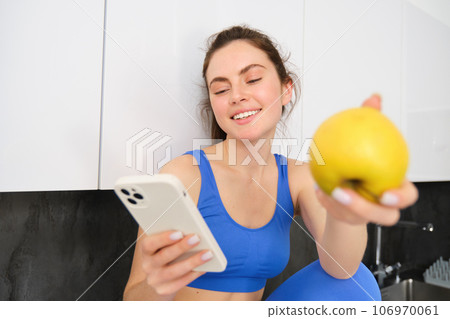 Portrait of smiling, happy fitness girl, sitting with an apple, laughing over smth on smartphone, browsing social media on mobile phone app 106970061