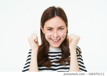 Image of young woman looking with pleased, satisfied face at camera, winning, shaking hands in triumph, celebrating victory, achievement, standing over white background 106970111