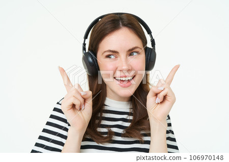 Dancing girl in wireless headphones pointing at empty space for music store promo text, showing advertisement, isolated over white background 106970148