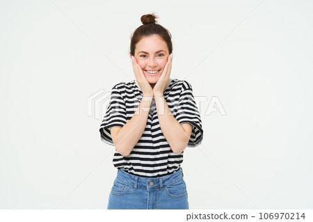 Portrait of cute smiling girl blushing, holds hands on cheeks and looks excited, receive happy news, stands over white background 106970214