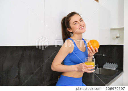 Indoor shot of woman after workout, standing in kitchen with fresh juice and an orange, drinking it 106970307