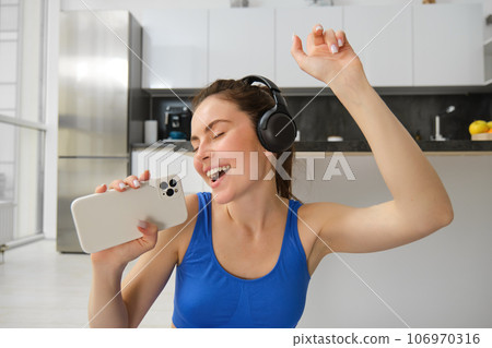 Happy young girl doing fitness, singing and having fun, listening music in headphones, holding smartphone, workout in living room 106970316