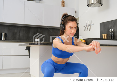 Image of young sportswoman, fitness instuctor doing squats workout at home, listening music in wireless headphones and stretching arms, doing exercises in living room 106970448