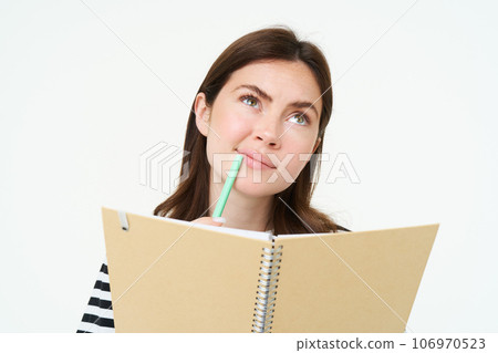Portrait of student doing her homework, holding notebook and pen, thinking while making notes in her notebook, standing over white background 106970523