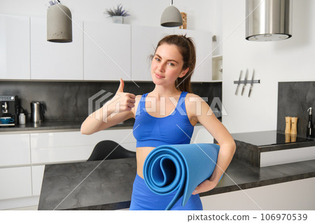 Attractive, sporty woman in blue activewear, shows thumb up, holds rubber yoga mat, workout at home, doing sport exercises in living room Attractive, sporty woman in blue activewear, shows thumb up, holds rubber yoga mat, workout at home, doing sport exercises in living room 106970539