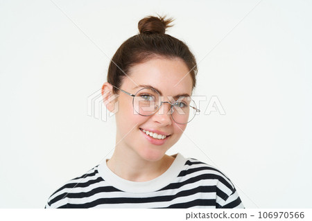 Close up portrait of cute young woman in glasses, looking at camera and smiling, trying on new spectacles, standing over white background 106970566