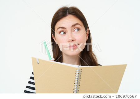 Portrait of brunette young woman, creative girl thinking, holding pen and notebook, making notes, writing in her diary, standing over white background 106970567