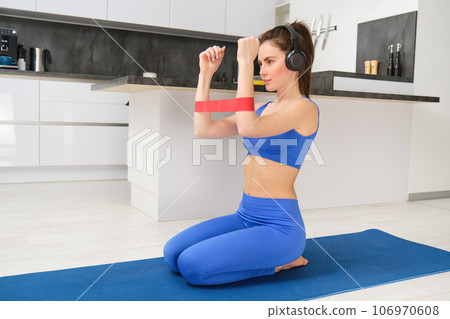 Young woman doing fitness workout at home, using elastic resistance band on arms, muscle exercises, sitting on rubber mat in living room 106970608