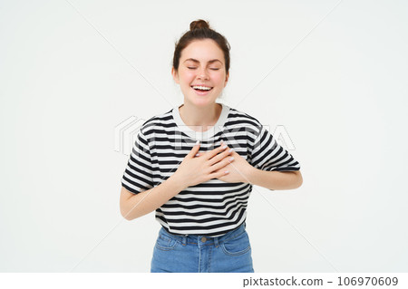 Portrait of happy, smiling young woman stands with eyes closed, holds hands on heart, laughs, remember something funny, isolated over white background 106970609
