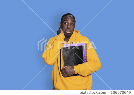 Young black man standing and holding notebooks on blue background while looking at camera frustrated Young black man standing and holding notebooks on blue background while looking at camera frustrated 106970649