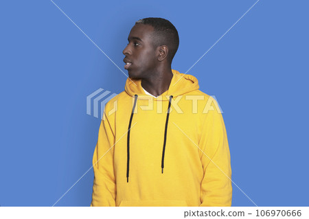 Portrait of cheerful young african-american guy wearing yellow hoodie sweater posing isolated on blue background. Carefree young millennial man looking aside Portrait of cheerful young african-american guy wearing yellow hoodie sweater posing isolated on blue background. Carefree young millennial man looking aside 106970666
