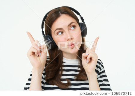 Portrait of young brunette woman in headphones, listening music in headphones, pointing fingers up, showing banner, advertisement, isolated over white background 106970711
