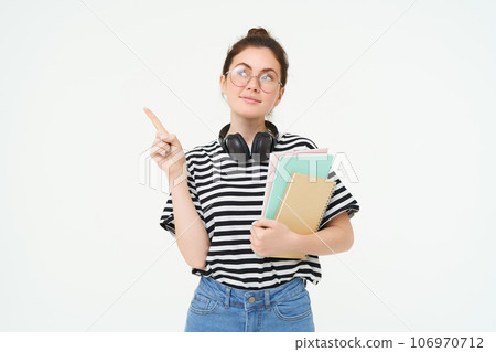 Image of smart young woman in glasses, student carries her notebooks and planners, points left with thinking face, making choice, deciding on smth, white background 106970712