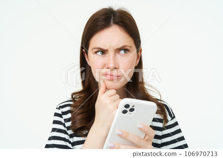 Portrait of girl with complicated face, holds smartphone and looks perplexed, thinks, makes difficult choice or decision, white background 106970713