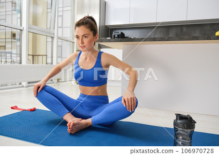 Indoor shot of active and healthy girl does workout from home, stretches her legs in yoga lotus pose, doing sports, fitness training in her house 106970728