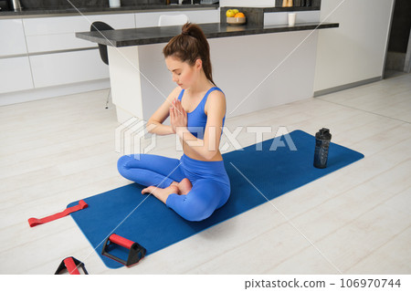 Fitness and sport concept. Young woman doing workout at home, sits on rubber yoga mat and stretching, wearing blue sports bra and leggings Fitness and sport concept. Young woman doing workout at home, sits on rubber yoga mat and stretching, wearing blue sports bra and leggings 106970744