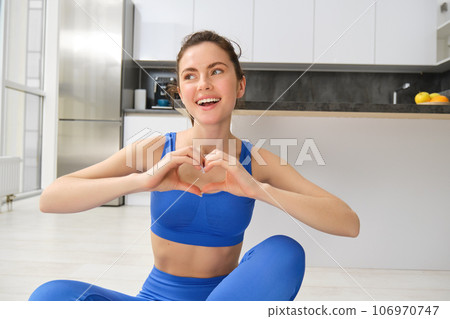 Portrait of beautiful, fit young woman in leggins and sportsbra, sits in lotus pose on yoga mat at home, shows heart sign and smiles at camera, workout, fitness instructor online video training Portrait of beautiful, fit young woman in leggins and sportsbra, sits in lotus pose on yoga mat at home, shows heart sign and smiles at camera, workout, fitness instructor online video training 106970747