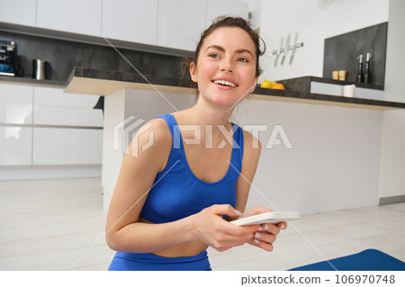 Portrait of healthy young sporswoman, smiling and holding smartphone, doing workout training at home in her house, fitness exercises 106970748