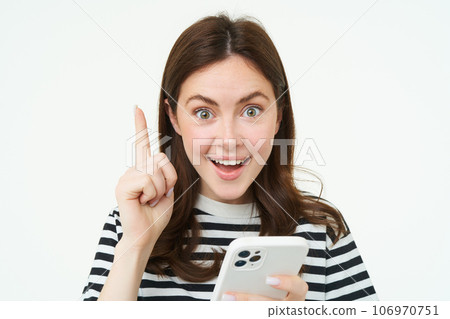 Got an idea. Smiling woman, raises her finger and holds smartphone, suggests something, makes eureka gesture, has a solution, white background 106970751