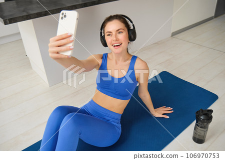 Portrait of fitness woman in wireless headphones, sits on fitness mat, doing selfies and photos during training exercises, working out at home Portrait of fitness woman in wireless headphones, sits on fitness mat, doing selfies and photos during training exercises, working out at home 106970753