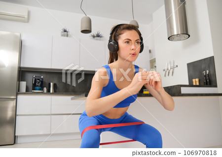 Image of beautiful fitness girl in headphones, does squats exercises with elastic band, resistance training, standing on yoga mat Image of beautiful fitness girl in headphones, does squats exercises with elastic band, resistance training, standing on yoga mat 106970818