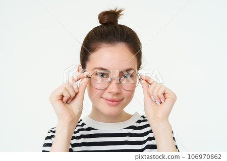Close up portrait of smiling, attractive young woman wearing new eyewear, trying on glasses for vision, standing over white background Close up portrait of smiling, attractive young woman wearing new eyewear, trying on glasses for vision, standing over white background 106970862