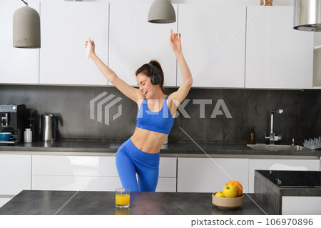 Portrait of carefree sportswoman, dancing in kitchen with smartphone, listening music in headphones, feeling energized and excited after workout 106970896