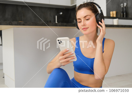 Portrait of woman in headphones, looking at smartphone, sitting on rubber mat and choosing music, doing yoga, workout training 106971030