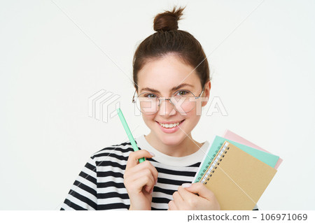 Close up portrait of young woman, tutor, online teacher in glasses, holding pen and documents, carries notebooks, stands over white background 106971069