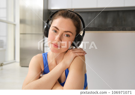 Close up portrait of brunette fitness girl, listens music in wireless headphones, smiles at camera, does workout alone at home, sits on floor and exercises 106971071