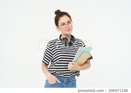 Education concept. Smiling brunette girl, student in casual clothes, holds her books, study material, wears headphones over neck, looks confident and happy, isolated over white background Education concept. Smiling brunette girl, student in casual clothes, holds her books, study material, wears headphones over neck, looks confident and happy, isolated over white background 106971106
