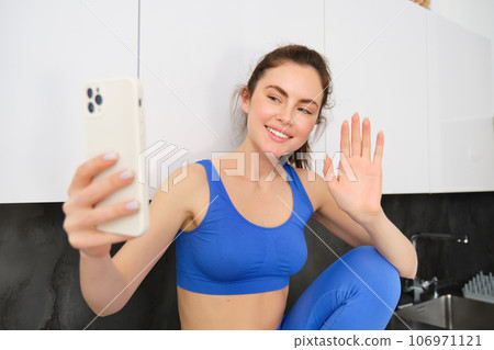 Portrait of young fitness girl, social media blogger, live streaming, saying hello at smartphone, waving hand and smiling, wearing activewear 106971121