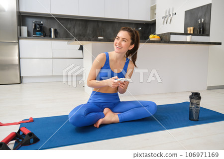 Portrait of woman using fitness app, doing workout from home, sits on yoga mat with smartphone in living room 106971169