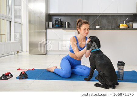 Image of young woman does workout from home, sits on yoga mat in living room and plays with her black dog, puppy distracts girl from doing exercises 106971170