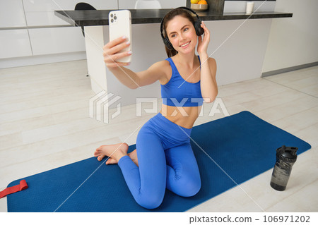 Portrait of fitness woman in wireless headphones, sits on fitness mat, doing selfies and photos during training exercises, working out at home Portrait of fitness woman in wireless headphones, sits on fitness mat, doing selfies and photos during training exercises, working out at home 106971202
