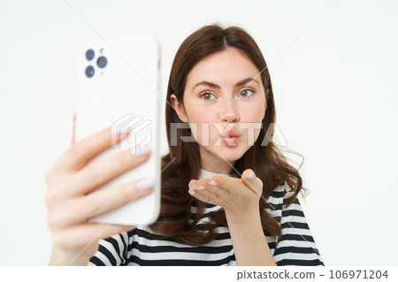 Portrait of young european woman taking selfie on smartphone, holding white mobile phone and posing for photos, isolated against white background 106971204