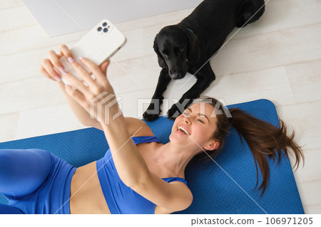 Portrait of happy young fitness woman, lying on mat and taking selfie with a dog in living room, workout with her pet Portrait of happy young fitness woman, lying on mat and taking selfie with a dog in living room, workout with her pet 106971205