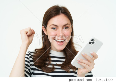 Enthusiastic brunette girl winning, holding smartphone and cheering, celebrating great news, standing with excited face expression, white background Enthusiastic brunette girl winning, holding smartphone and cheering, celebrating great news, standing with excited face expression, white background 106971222