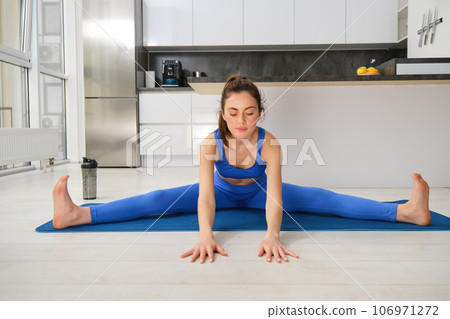 Portrait of young woman does gymnastics training at home, workout, making splits on rubber fitness mat, sitting on floor with feet stretched sideways Portrait of young woman does gymnastics training at home, workout, making splits on rubber fitness mat, sitting on floor with feet stretched sideways 106971272