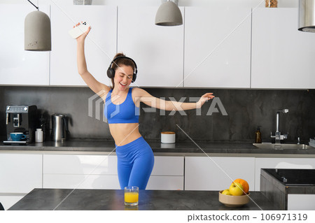 Portrait of beautiful sports woman, wearing activewear, dancing in kitchen with headphones, listening music and having fun, feeling energy after workout session 106971319