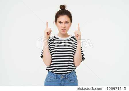 Portrait of shocked woman, raise eyebrows and points fingers up, looks complicated, puzzled by something, standing over white background 106971345